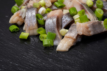 Slices of Salted Boneless wild-caught Atlantic Herring garnished with Chopped Green Onion Scallions. Black natural stone background. Clupea harengus. 