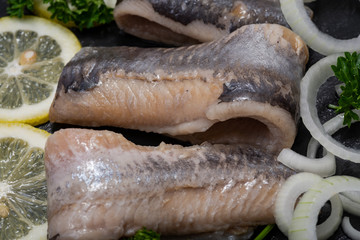 Salted Deboned Herring Fillets garnished with Yellow Onion, Lemon and Fresh Parsley. Natural black stone background. Clupea harengus.