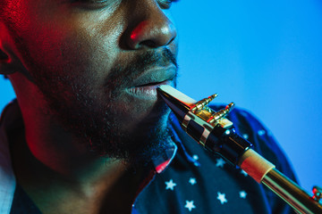 Young african-american jazz musician playing the saxophone on blue studio background in trendy neon light. Concept of music, hobby. Joyful attractive guy improvising. Retro colorful portrait of artist