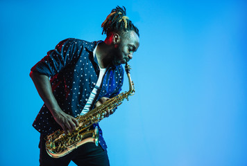 Young african-american jazz musician playing the saxophone on blue studio background in trendy neon...