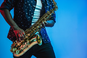 Young african-american jazz musician playing the saxophone on blue studio background in trendy neon light. Concept of music, hobby. Joyful attractive guy improvising. Retro colorful portrait of artist - obrazy, fototapety, plakaty