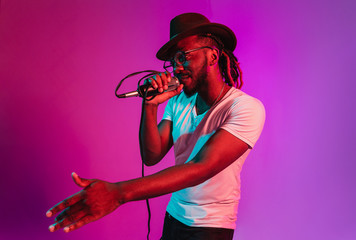 Young african-american jazz musician with microphone singing a song on purple studio background in trendy neon light. Concept of music, hobby, inspirness. Colorful portrait of joyful attractive artist