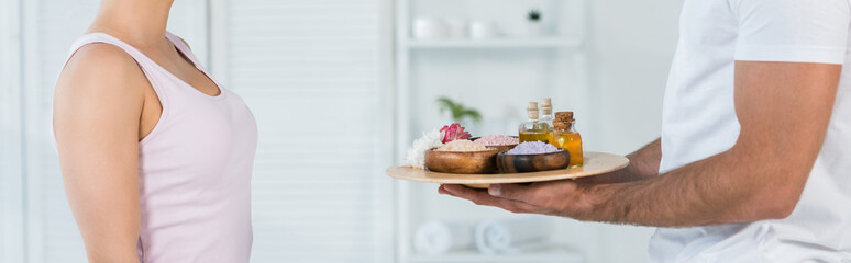 panoramic shot of  masseur holding tray with sea salt and oil in bottles near woman