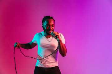 Young african-american jazz musician with microphone singing a song on purple studio background in trendy neon light. Concept of music, hobby, inspirness. Colorful portrait of joyful attractive artist