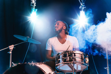 Young african-american jazz musician or drummer playing drums on blue studio background in glowing...