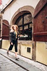 The girl walks with a camera in Prague. A young blonde woman walks along a narrow cobbled street in Prague, Czech Republic. Stylish blonde girl in a leather jacket.