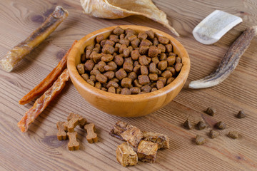 dried dog food in a bowl and different snack, chicken filet, antlers, lung, ear on wooden background