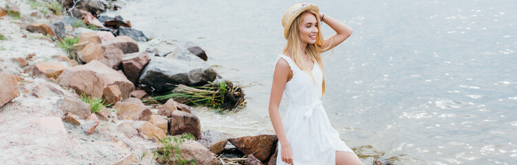 panoramic shot of cheerful young blonde woman touching straw hat and looking at sea