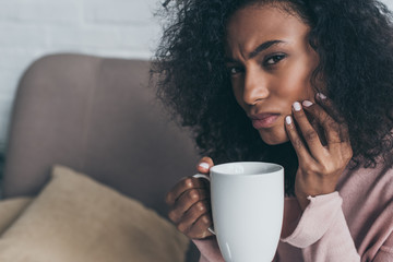 unhappy african american woman looking at camera while suffering from tooth pain and holding coffee...