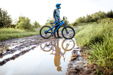 Fototapeta na wymiar Professional well-equipped cyclist crossing mountain dirt road with puddles during the sunset