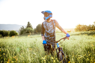 Portrait of a professional well-equipped cyclist walking with bicycle on the beautiful green field on the mountains during the sunset