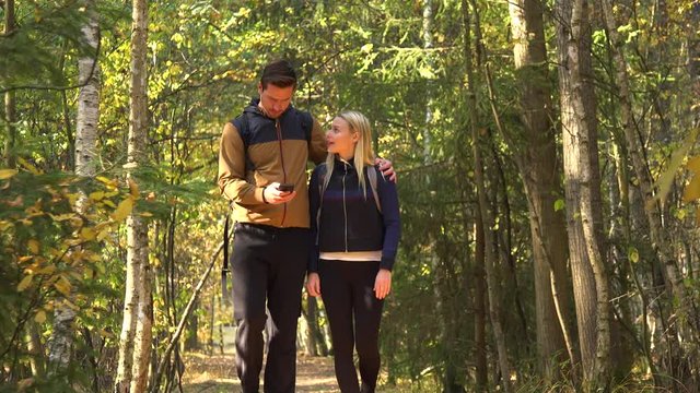 A hiking couple walks through a forest on a sunny day and looks at a smartphone