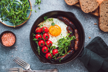 Breakfast time, fried egg with sausages and cherry tomatoes in a black iron pan, served microgreens.