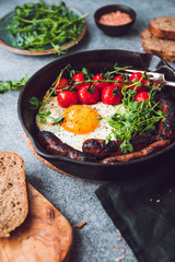Breakfast time, fried egg with sausages and cherry tomatoes in a black iron pan, served microgreens.