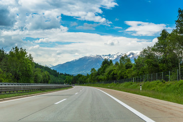 Fototapeta premium Autobahn or highway in the mountains with clear marking surrounded by vibrant green trees under blue sky. Stunning view and a snow-covered mountain in the background. The Alps, Austria