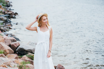 happy young blonde woman touching straw hat and looking at sea