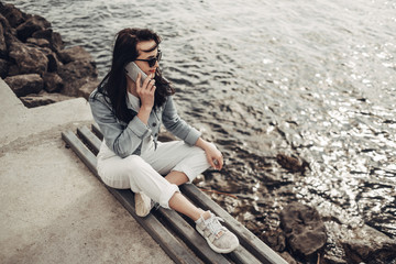 Beautiful Brunette Girl in Sunglasses White Pants and Jeans Jacket Near the Lake