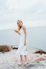 happy blonde girl in white dress smiling while standing on beach