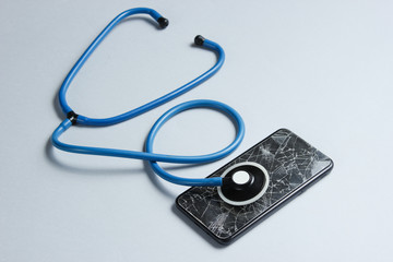 The concept of service assistance in the breakdown of  smartphone. Broken glass screen of a smartphone, stethoscope on gray background