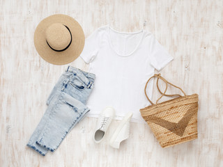 White t-shirt, light blue ripped jeans, white sneakers, wicker bag, straw boater hat on white wooden background. Overhead view of woman's casual day outfit. Trendy spring or summer look. Flat lay.