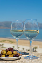 Two wine glasses with white wine served with olives on outdoor terrace witn blue sea and mountains view on background