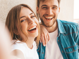 Smiling beautiful girl and her handsome boyfriend in casual summer clothes. Happy family taking selfie self portrait of themselves on smartphone camera. Having fun on the street background - obrazy, fototapety, plakaty