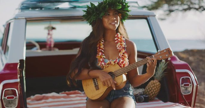 Portrait of beautiful young woman singing smiling and playing the ukulele, sitting in a vintage beach cruiser car at sunset, happy woman wearing flower lei and tiara, hawaiian island cruising