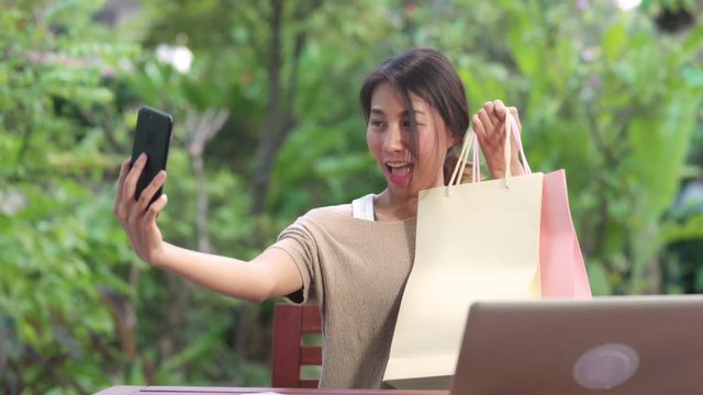 Asian woman using mobile phone selfie post in social media, female relax feeling happy showing shopping bags sitting on table in the garden in morning. Lifestyle women relax at home concept.