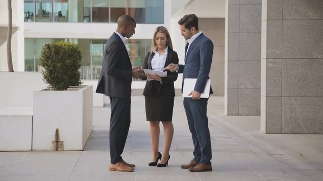 Young business colleagues discussing papers outdoor. Professional multiethnic business people with documents and laptop standing outside modern office building. Business concept