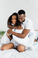 african american man hugging his charming girlfriend, sitting on white bed