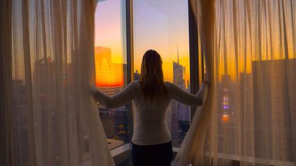 CLOSE UP: Unrecognizable woman opens the curtains and looks at city at sunset.