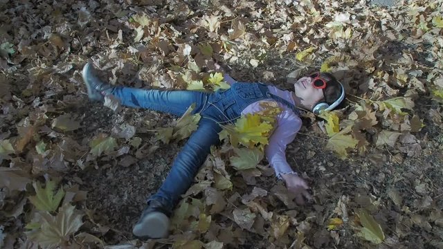 Kid playing snow angel among fallen leafs. Happy little girl enjoys music with headphones lying on autumn leaves.