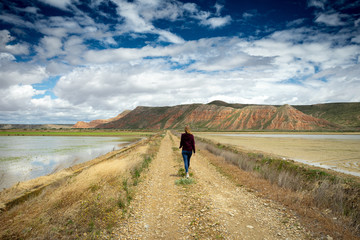 une femme de dos marche sur un chemin entouré de champs inondés en direction de montagnes ocres