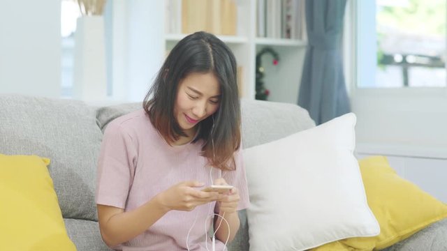 Asian woman listening music and using smart phone, female using relax time lying on home sofa in living room at home. Happy female listening music with headphones concept.