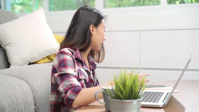 Freelance Asian woman working at home, business female working on laptop and using mobile phone talking with customer on sofa in living room at home. Lifestyle women working at home concept.