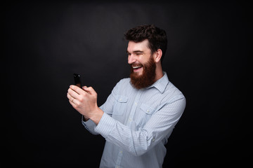 Handsome cheerful bearded man using mobile, typing sms, standing over dark isolated background