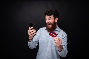 Cheerful bearded man using a mobile phone, and holding a credit card, over dark isolated background