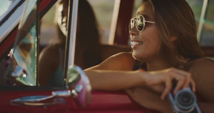 Young attractive woman sitting and smiling in a red vintage beach cruiser car at the beach looking out at the ocean with aviators on at sunset, hawaiian island surf concept, friends at the beach