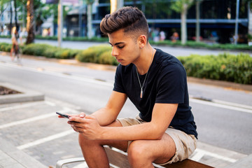 Fototapeta premium Young fashionable handsome man on the street of modern city chatting and typing on a cell phone. Communication and messenger concept