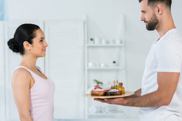 handsome masseur holding tray with sea salt and oil in bottles near attractive woman