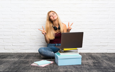 Teenager girl sitting on the floor with her laptop smiling and showing victory sign