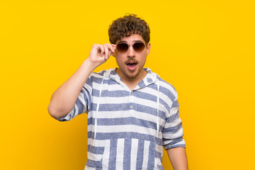 Blonde man over yellow wall with glasses and surprised