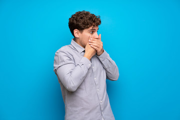 Blonde man over blue wall covering mouth and looking to the side