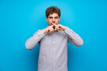 Blonde man over blue wall showing a sign of silence gesture
