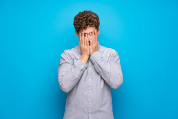 Blonde man over blue wall with tired and sick expression