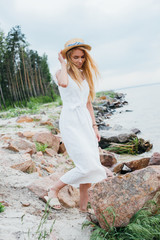 beautiful blonde woman standing and touching straw hat near sea