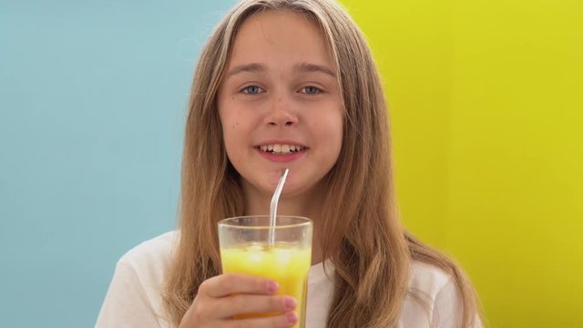 Close-up of a pretty teen girl on a yellow-blue background drinking orange juice with ice through a straw from a large glass cup