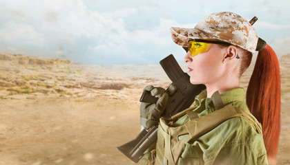 Portrait of young and beautiful military soldier woman with red hair holding an automatic rifle M16 on desert background with space for text.