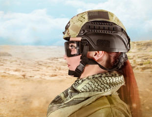 Portrait of fully equipped young military soldier woman with red hair in helmet on desert background, side view.