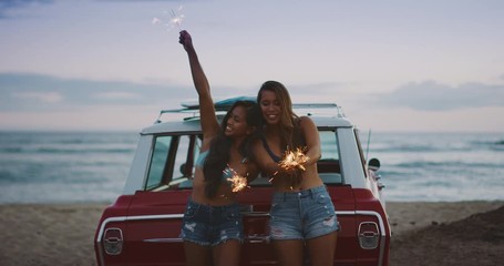 Two attractive hipster surf girls playing with sparkler fireworks next to a vintage car at the beach, summer fun at the beach